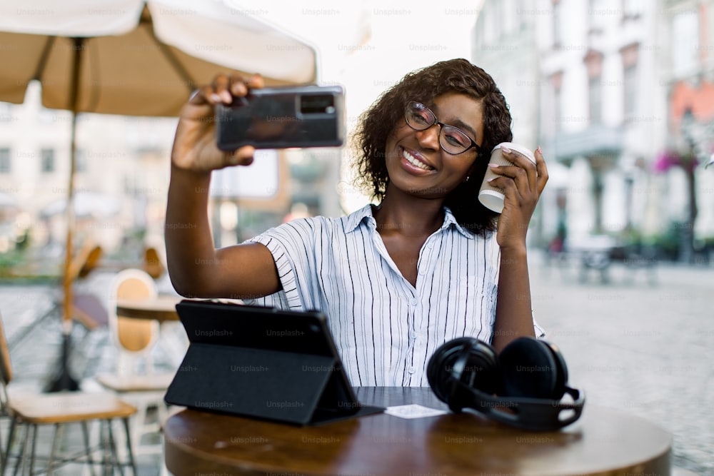 Encantadora joven africana de piel oscura con camisa a rayas, haciendo una foto selfie o haciendo una videollamada, mientras está sentada en un café de la ciudad al aire libre, bebiendo café y usando una tableta digital.