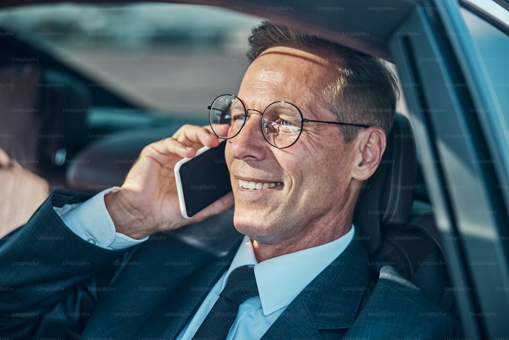 Jolly Geschäftsmann in elegantem Anzug und Brille sitzt im Transport und plaudert auf dem Handy