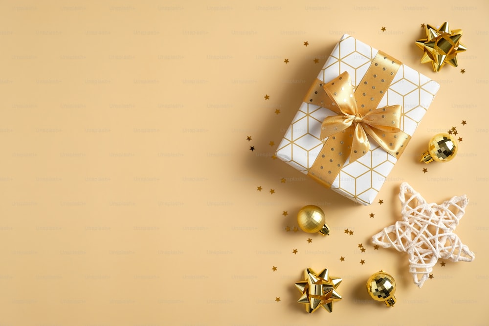 Luxury Christmas composition. Gift box with golden ribbon bow, balls decorations, star, confetti on yellow background. Flat lay, top view, copy space. Season greeting card mockup.