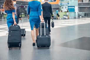 Des agents de bord en uniforme bleu et des pilotes avec des sacs à bagages à roulettes marchant dans le terminal de l’aéroport