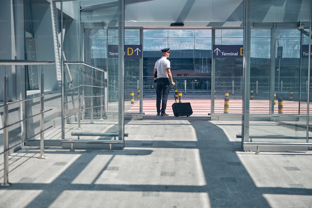 Employé d’une compagnie aérienne avec un sac à bagages à roulettes debout près de la porte de sortie du terminal de l’aéroport