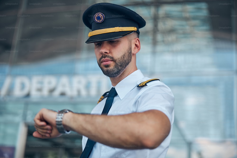 Good-looking young man airline captain in pilot hat checking time before the flight at airport