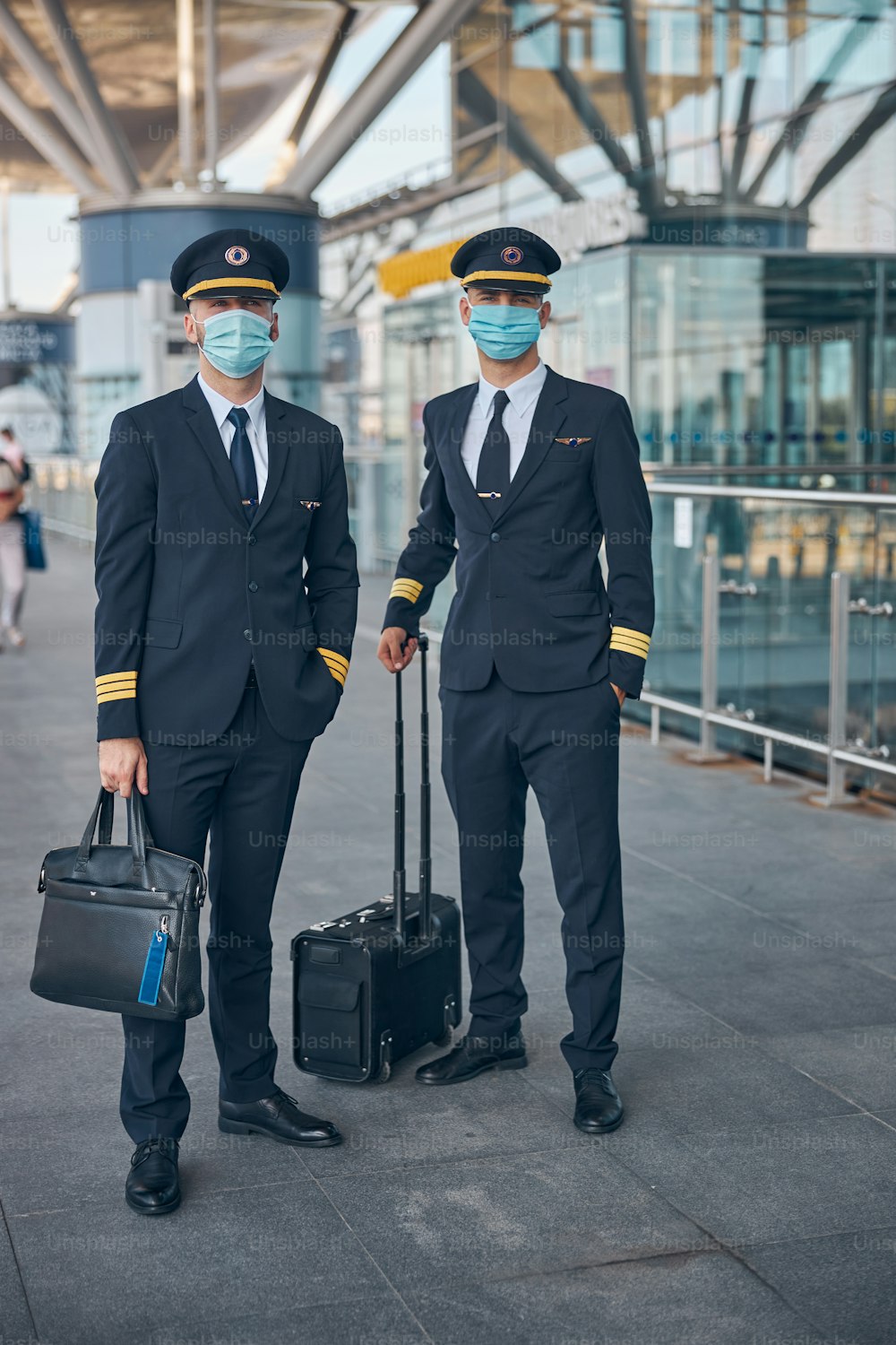 Hübsche junge Piloten in Schutzmasken, die Reisetaschen halten und in die Kamera schauen, während sie am Flughafen auf ihren Flug warten