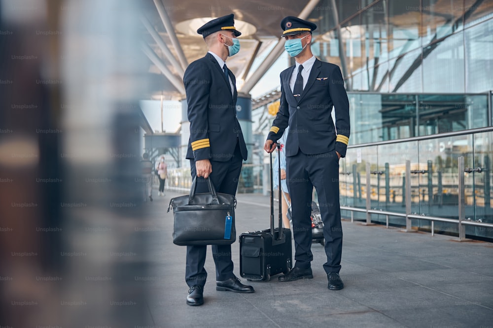 Dos jóvenes guapos trabajadores de una aerolínea con máscaras protectoras esperan el vuelo en el aeropuerto