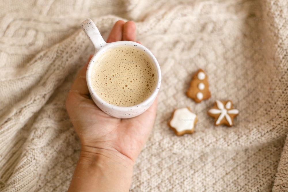 Hand holding warm coffee on background of cozy knitted sweater with christmas gingerbread cookies. Atmospheric winter hygge. Happy Holidays!