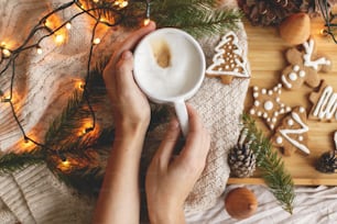 Hands holding warm coffee on background of christmas gingerbread cookies, cozy knitted sweater, fir branches with pine cones and lights. Hello winter. Merry Christmas and Happy Holidays!