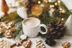 Hello winter, cozy moody image. Hot coffee in stylish white cup with homemade gingerbread cookies and pine cones decorations, fir branches and warm lights on white wooden table