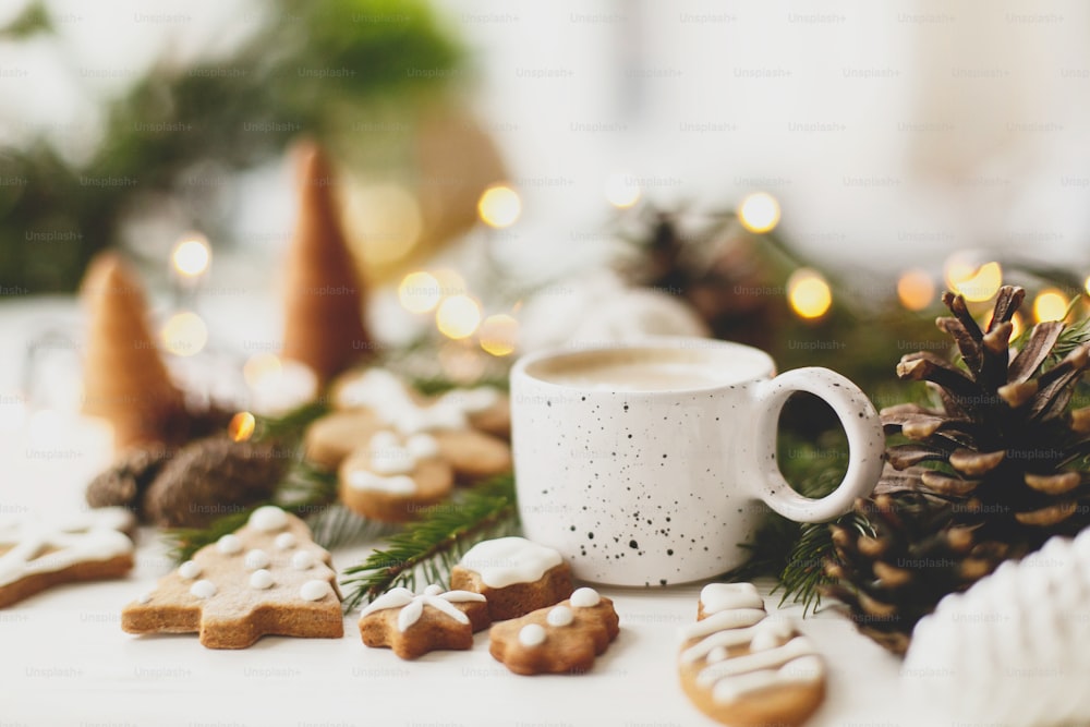 Christmas gingerbread cookies, coffee in stylish white cup, pine cones  and warm lights on white wooden table. Hello winter, cozy moody image with selective focus