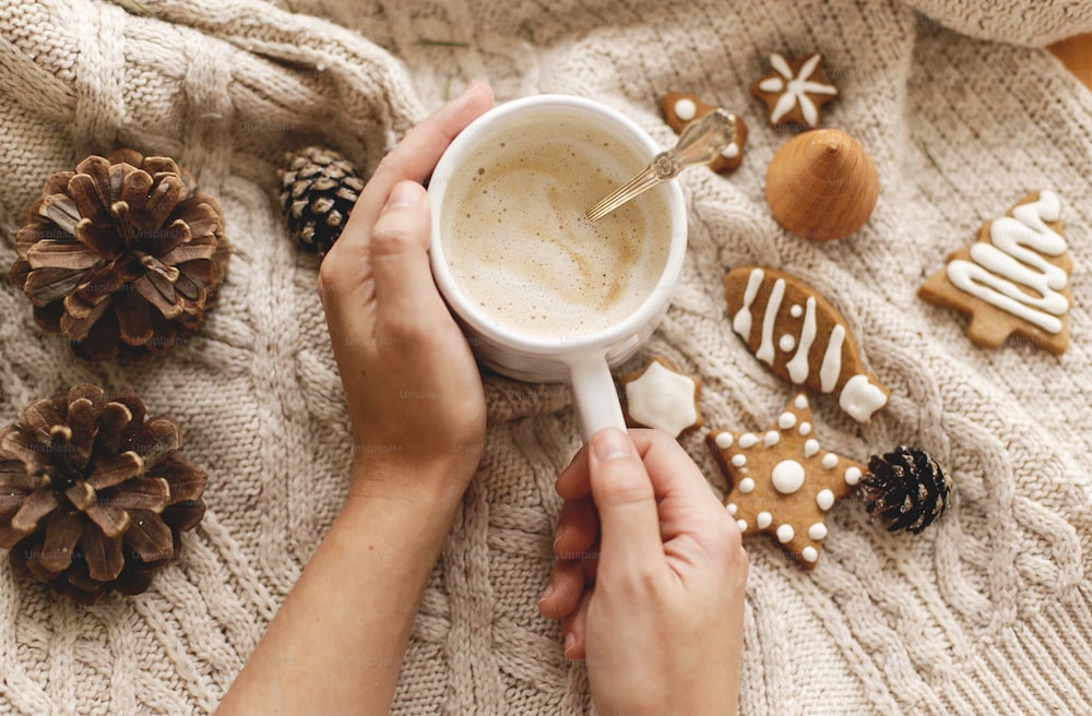 Hands holding warm coffee on background of cozy knitted sweater with christmas gingerbread cookies and pine cones, flat lay. Atmospheric winter hygge. Happy Holidays!