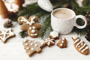 クリスマスのジンジャーブレッドクッキー、スタイリッシュな白いカップに入ったコーヒー、白い木製のテーブルに松ぼっくりとモミの枝。こんにちは冬、居心地の良い不機嫌なイメージ