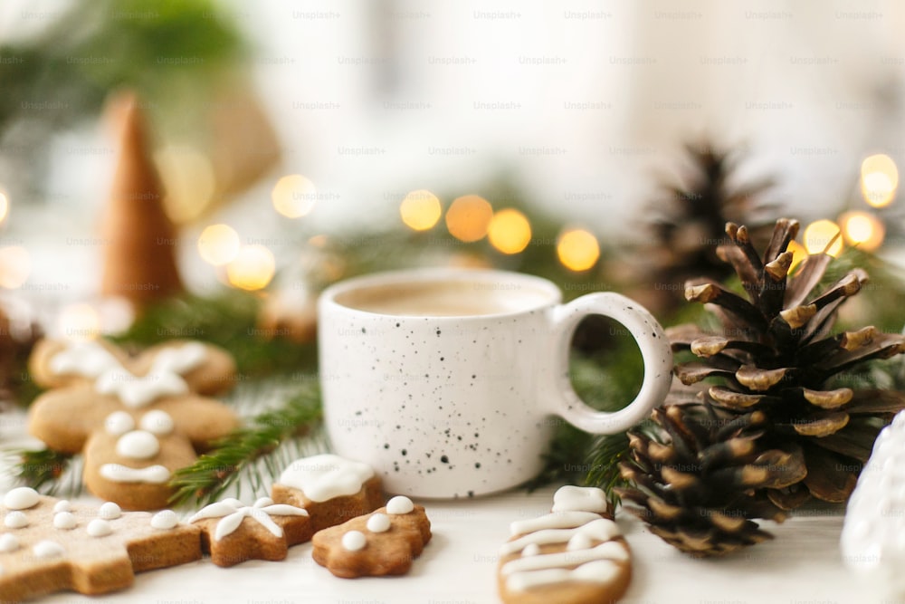 Hot coffee in stylish white cup with homemade gingerbread cookies and pine cones decorations, fir branches and warm lights on white wooden table. Hello winter, cozy moody image