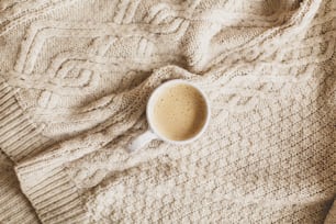 Warm coffee in stylish cup on cozy knitted sweater, top view. Atmospheric winter hygge or cozy autumn flat lay. Happy Holidays