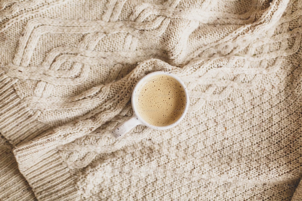 Warm coffee in stylish cup on cozy knitted sweater, top view. Atmospheric winter hygge or cozy autumn flat lay. Happy Holidays