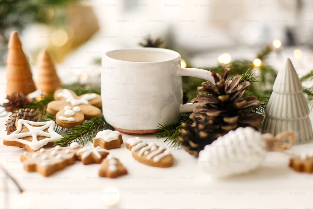 Christmas gingerbread cookies, coffee in stylish white cup, pine cones  and warm lights on white wooden table. Hello winter, cozy moody image