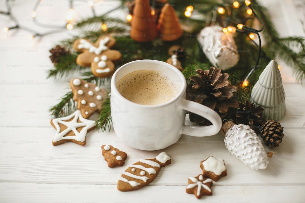 Café quente em elegante xícara branca com biscoitos caseiros de gengibre e decorações de pinhas, ramos de abeto e luzes quentes na mesa de madeira branca. Olá inverno, imagem aconchegante e mal-humorada