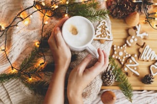 Hands holding warm coffee on background of christmas gingerbread cookies, cozy knitted sweater, fir branches with pine cones and lights. Hello winter, atmospheric image. Happy Holidays!