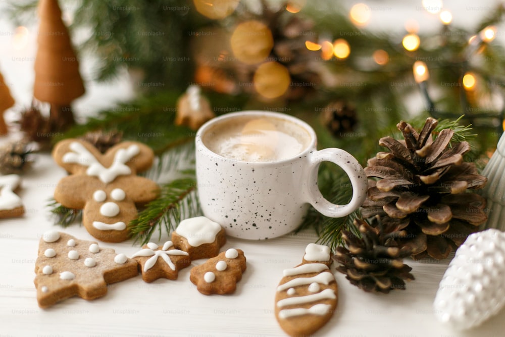 Happy Holidays! Christmas gingerbread cookies, coffee in stylish white cup, pine cones  and warm lights on white wooden table. Hello winter, cozy moody image