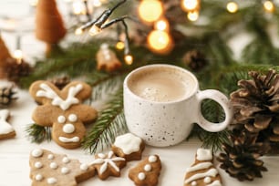 Happy Holidays! Christmas gingerbread cookies, coffee in stylish white cup, pine cones  and warm lights on white wooden table. Hello winter, cozy moody image