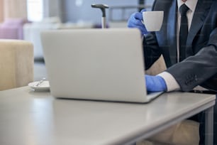 Cropped head of male in suit and latex gloves using notebook and drinking coffee before flight