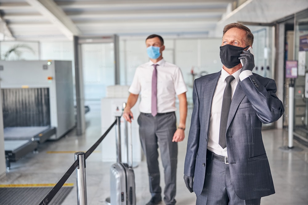 Mature elegant male in mask is making call while assistant is carrying luggage before departure