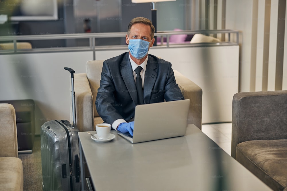 Top view waist up portrait of elegant man in suit wearing protective mask and gloves while working on notebook before flight