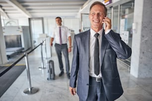 Fröhlicher, eleganter Mann in Krawatte geht am Flughafen und spricht auf dem Smartphone, während der Leibwächter Gepäck trägt
