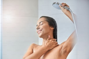 Affascinante signora con gli occhi chiusi che versa acqua sui capelli e sorride mentre usa il soffione della doccia a mano