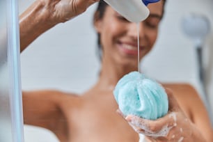 Close up of charming lady holding exfoliating bath sponge and smiling while taking shower at home