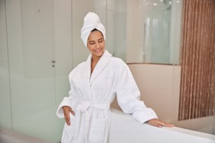 Attractive lady with towel on her head wearing white soft bathrobe while looking down and smiling