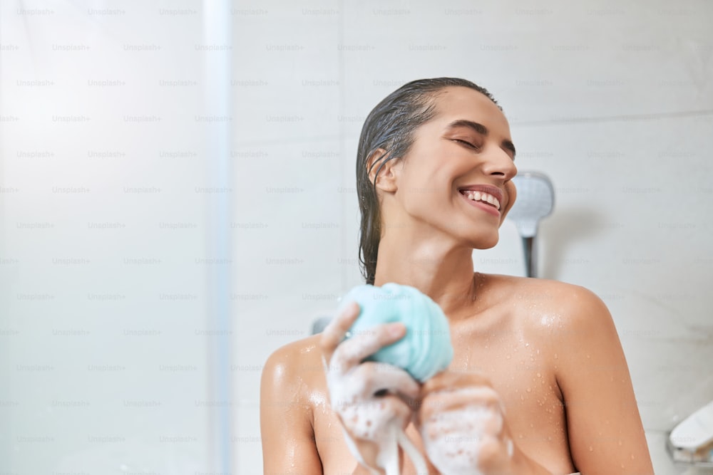 Charming lady with bath washcloth in her hands closing eyes and smiling while taking shower at home