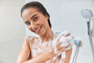 Attractive lady with foam on her skin looking at camera and smiling while washing body with bath sponge
