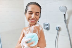 Cheerful lady with foam on her skin looking down and smiling while washing body with exfoliating sponge