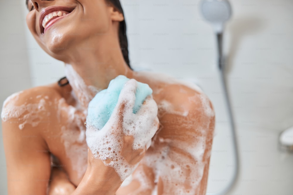 Close up of cheerful lady with foam on her skin washing body with exfoliating sponge