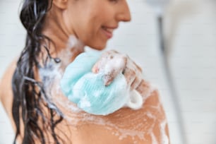Close up of cheerful lady with foam on her skin cleaning shoulder with bath sponge