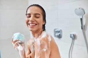 Beautiful joyful lady with exfoliating sponge in her hand taking shower at home