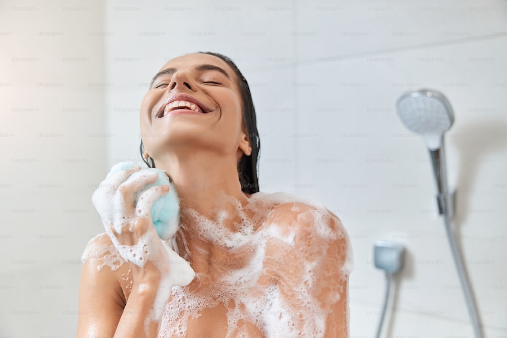 Signora graziosa sorridente con schiuma sulla pelle che usa una spugna da bagno esfoliante mentre fa la doccia a casa