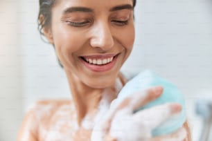 Primer plano de una linda dama sonriente usando una esponja de baño exfoliante mientras se ducha en casa
