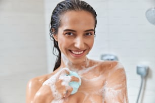 Beautiful lady with foam on her skin looking at camera and smiling while washing body with exfoliating sponge