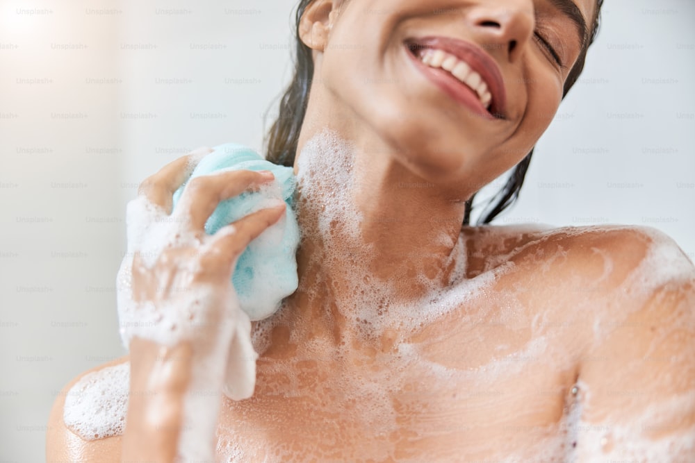 Close up of smiling lady with foam on her skin washing body with exfoliating sponge
