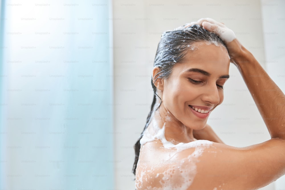 Beautiful lady with foam on her skin looking down and smiling while taking shower at home