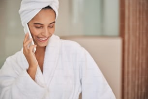 Attractive lady in white bathrobe having phone conversation and smiling