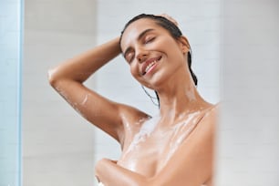 Pretty lady with foam on her skin touching her wet hair and smiling while taking shower at home