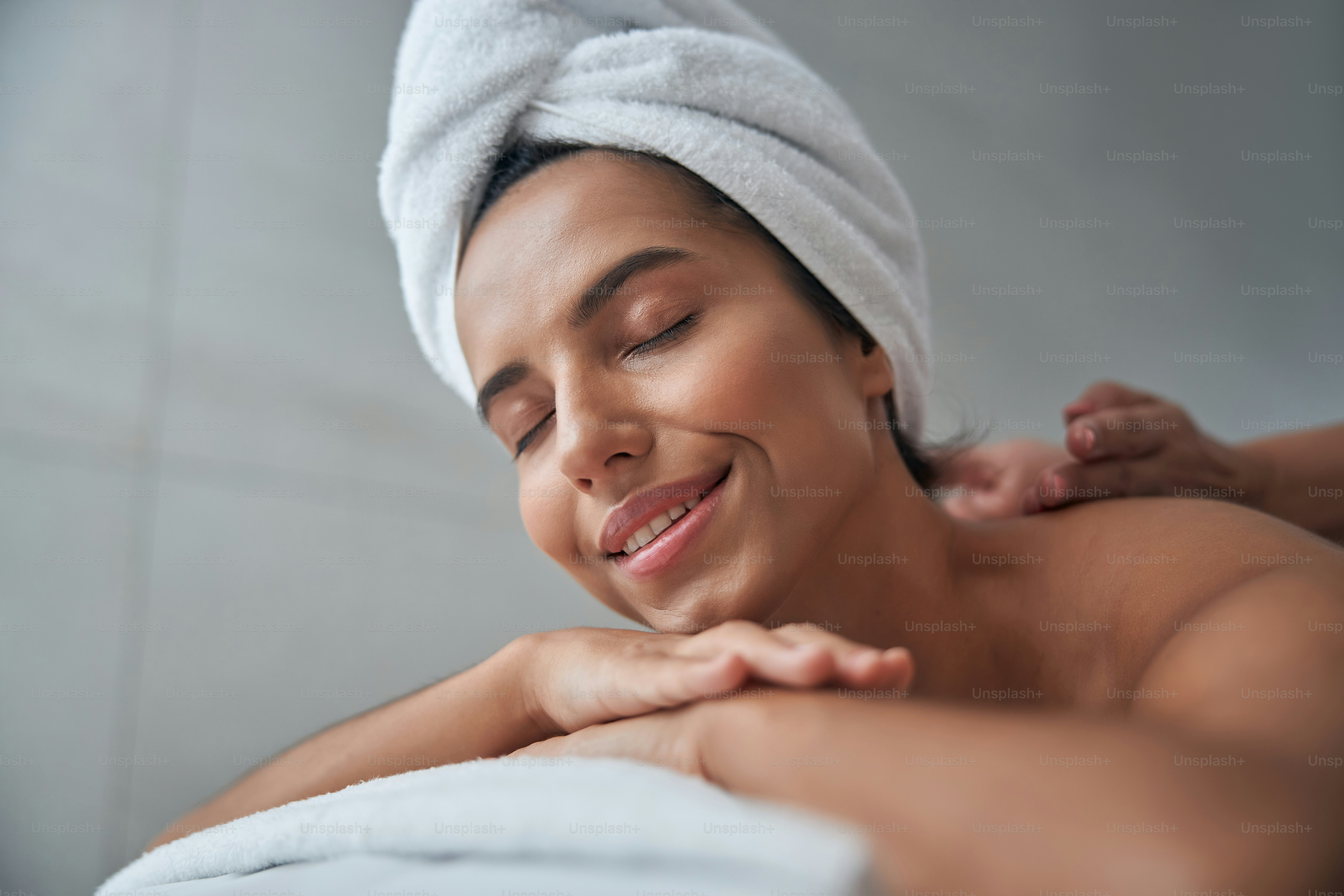 Wanita cantik dengan handuk mandi di kepalanya berbaring di atas meja pijat dan tersenyum sambil menerima pijat punggung profesional