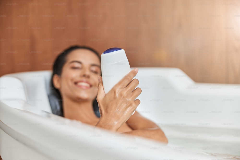 Primer plano de una señora sonriente con una botella de gel de baño en la mano relajándose en la bañera en casa