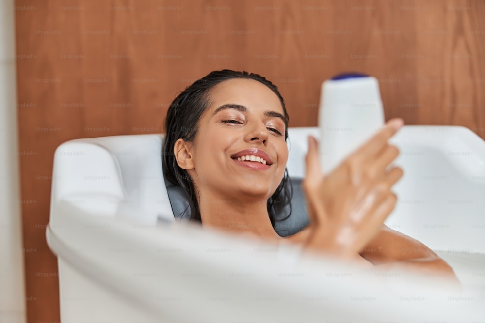 Charming lady with wet hair looking at body wash and smiling while relaxing in bathtub at home