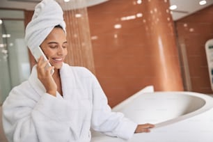 Beautiful lady in white bathrobe having phone conversation and smiling while standing by bathtub in spa salon