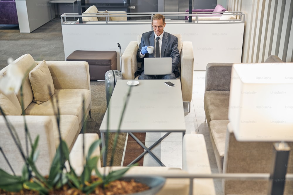 Draufsicht des fröhlichen eleganten Geschäftsmannes, der in Gummihandschuhen im Flughafen sitzt, Kaffee trinkt und Notebook benutzt