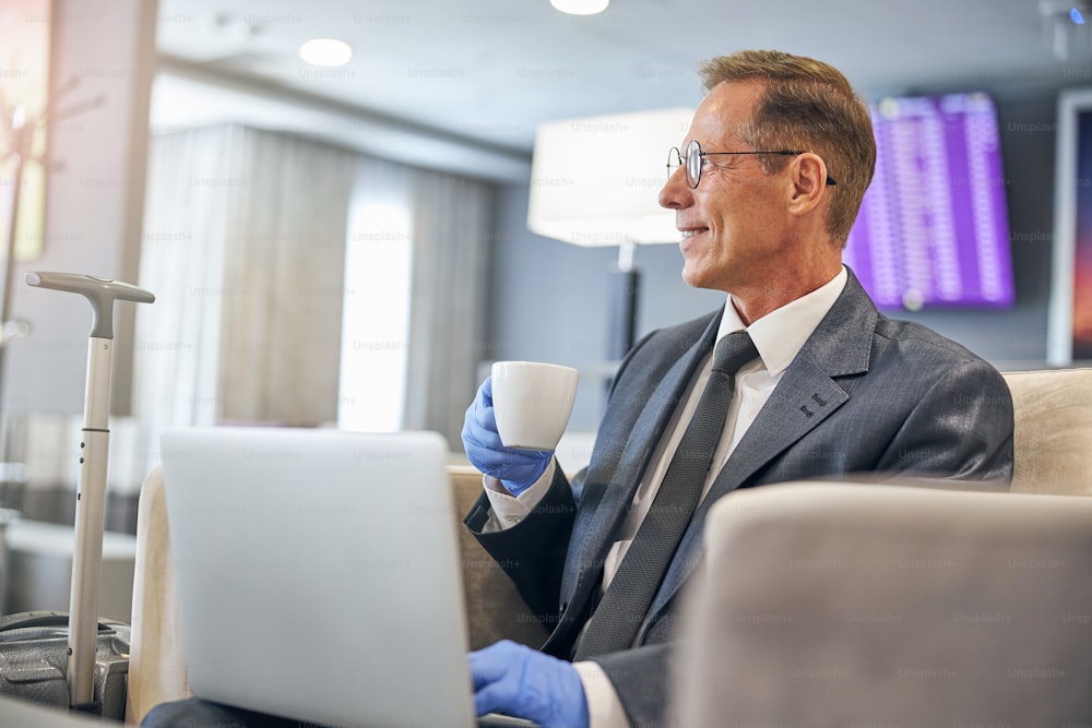 Lächelnder eleganter Mann mit Latexhandschuhen beim Kaffeetrinken mit Notebook vor dem Flug während der Pandemie