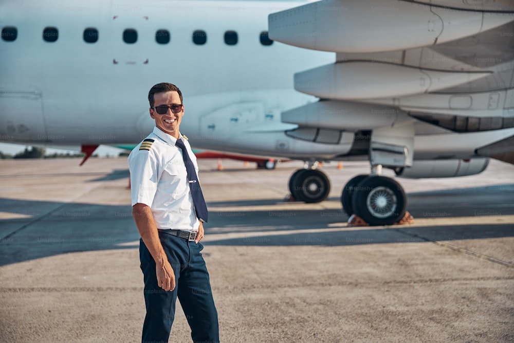 Ritratto dalla vita in su di un giovane allegro in uniforme e occhiali da sole in piedi vicino all'aereo dopo l'arrivo