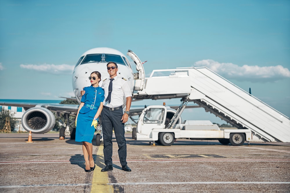 Full length portrait of joyful young stewardess and pilot standing together near passenger plane outdoors